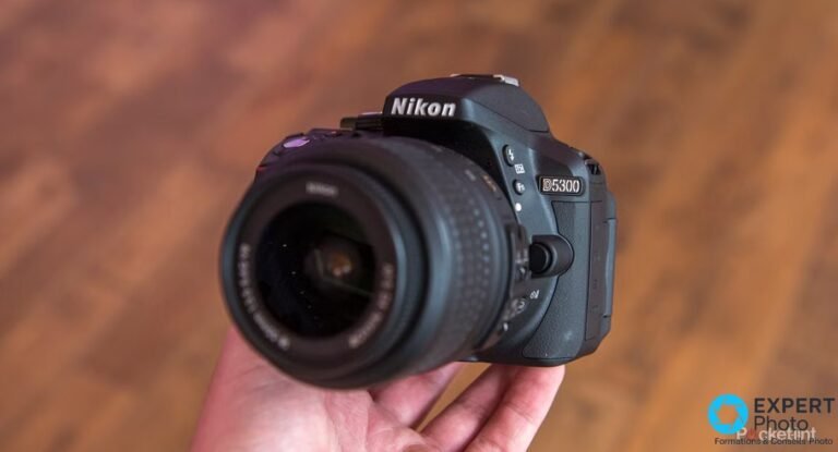 Nikon D5300 reflex tenu en main, vu de face avec un objectif Nikon monté sur la baïonnette du boîtier.