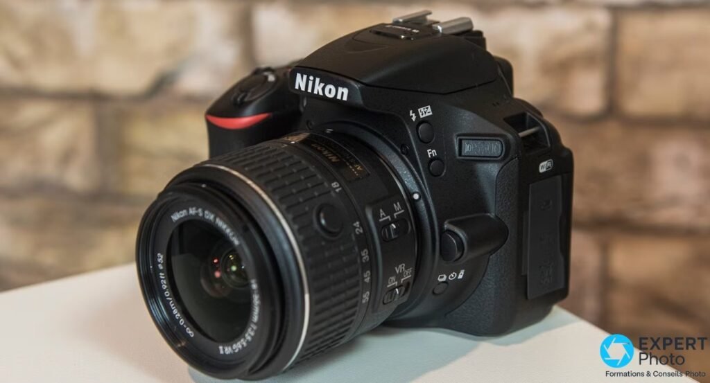 Nikon D5500 vu de trois quarts avant droite avec objectif AF-S DX 18-55 mm sur fond de mur de briques flou. 