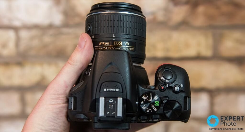Nikon D5500 tenu en main vu de dessus, molette de modes M/A/S/P/AUTO/SCENE/EFFECTS et microphone stéréo visibles.