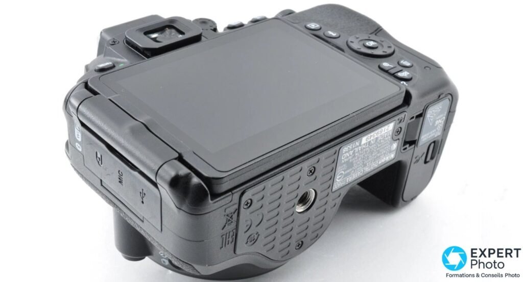 Nikon D5600 vu en 3/4 arrière dessous, écran fermé contre le boîtier, ports MIC et USB visibles côté gauche.