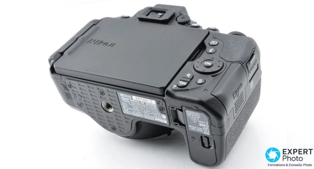 Nikon D5600 vu en 3/4 arrière dessus, écran orientable replié dos visible, étiquette de série visible sous le boîtier.