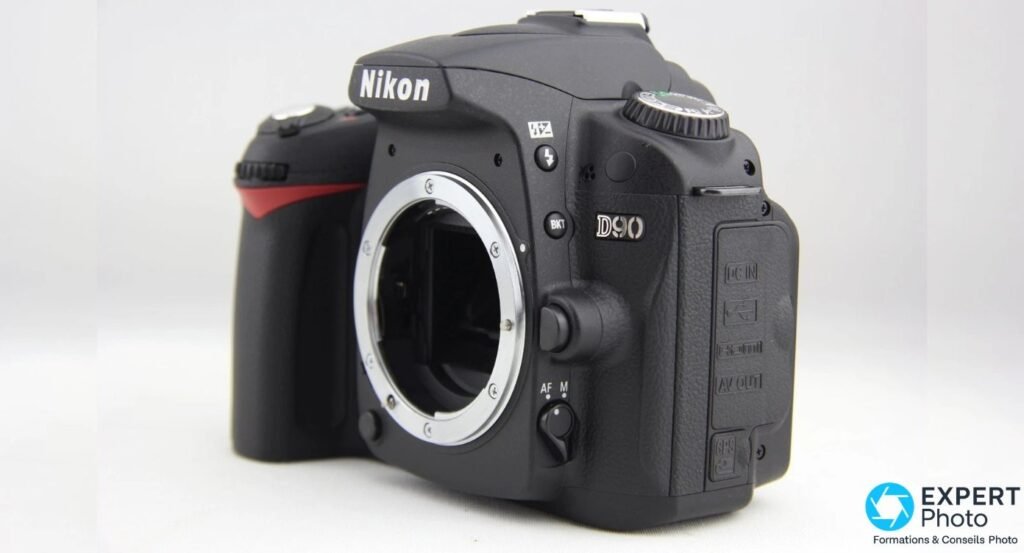 Boîtier nu Nikon D90 de trois quarts avant, monture Nikon F ouverte et sélecteur AF/M, fond clair