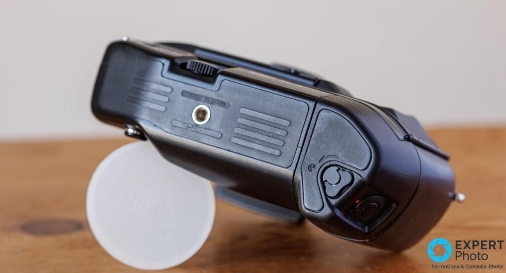 Vue du dessous d'un Nikon F4s, semelle du grip MB-21 avec écrou trépied, déclencheur vertical et cap Nikon posé à gauche