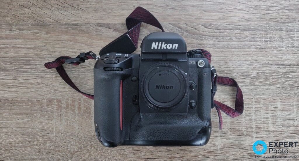 Nikon F5 vu de face sur parquet bois, couvre-boîtier Nikon en place, poignée verticale et sangle bordeaux étalée