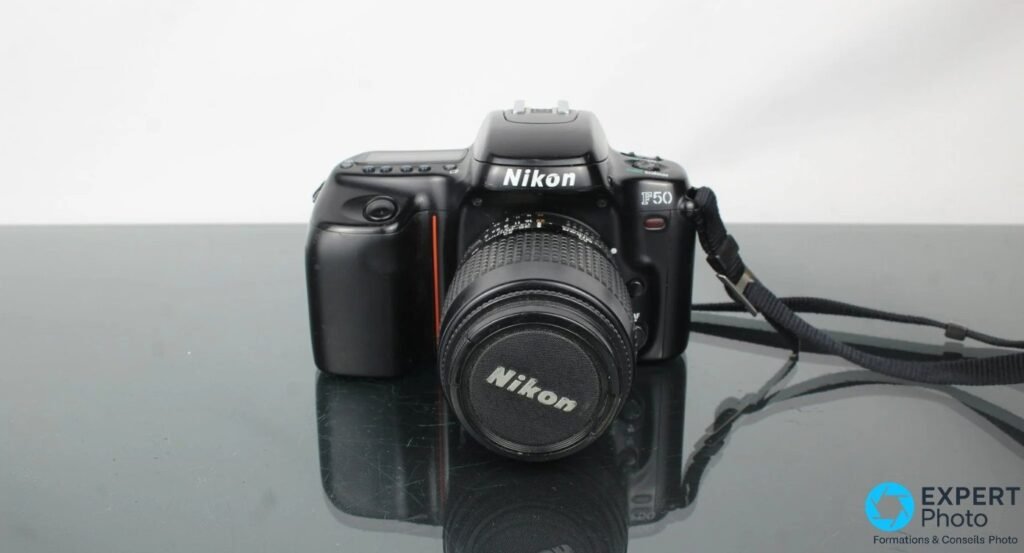 Boitier Nikon F50 vu de face, objectif Nikon monte bouchon en place, sangle noire, fond clair et table sombre.