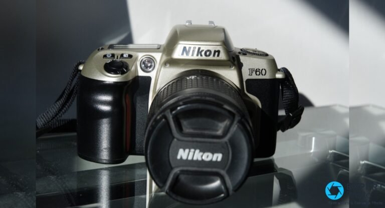 Nikon F60 couleur champagne vu de face, objectif zoom monté avec capuchon Nikon, courroie noire, sur surface réfléchissante