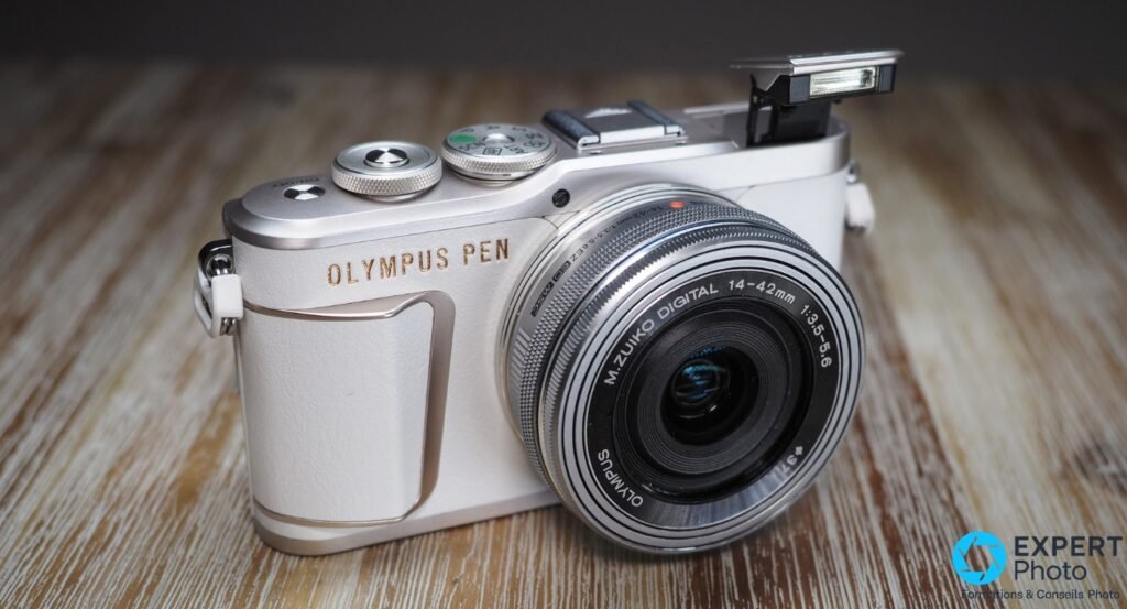 Olympus PEN E-PL10 blanc, vue 3/4 avant, objectif M.Zuiko 14-42 mm 1:3,5-5,6 monté, flash intégré relevé