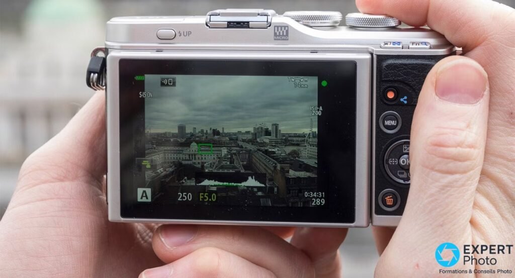 Olympus PEN E-PL9 vu de dos tenu à deux mains, écran arrière en Live View affichant une scène urbaine. 