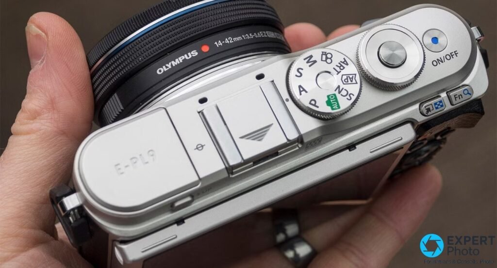 Olympus PEN E-PL9 vu de dessus, tenu en main, molette de modes et inscription E-PL9 visible.