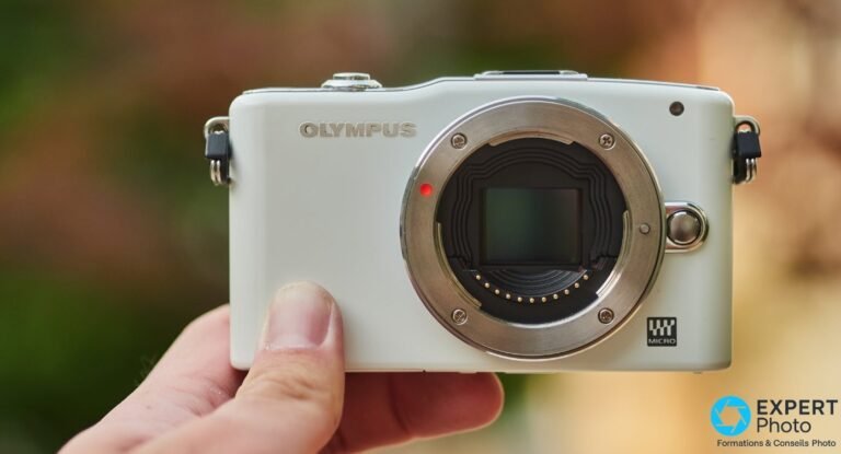 Olympus PEN E-PM1 finition blanche tenu en main, monture Micro Four Thirds ouverte montrant le capteur, fond extérieur flou