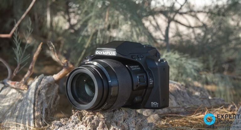Olympus Stylus SP-100EE, bridge superzoom noir vu de trois quarts avant, objectif déployé, logo OLYMPUS sur le capot.
