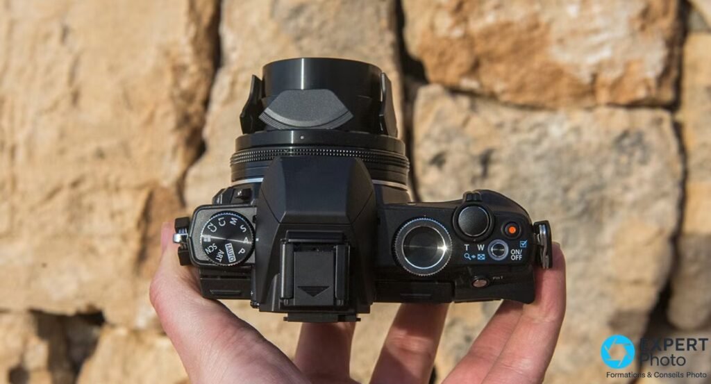  Olympus Stylus 1 vu du dessus tenu dans une main, format compact bridge avec molette de modes et objectif zoom déployé. 