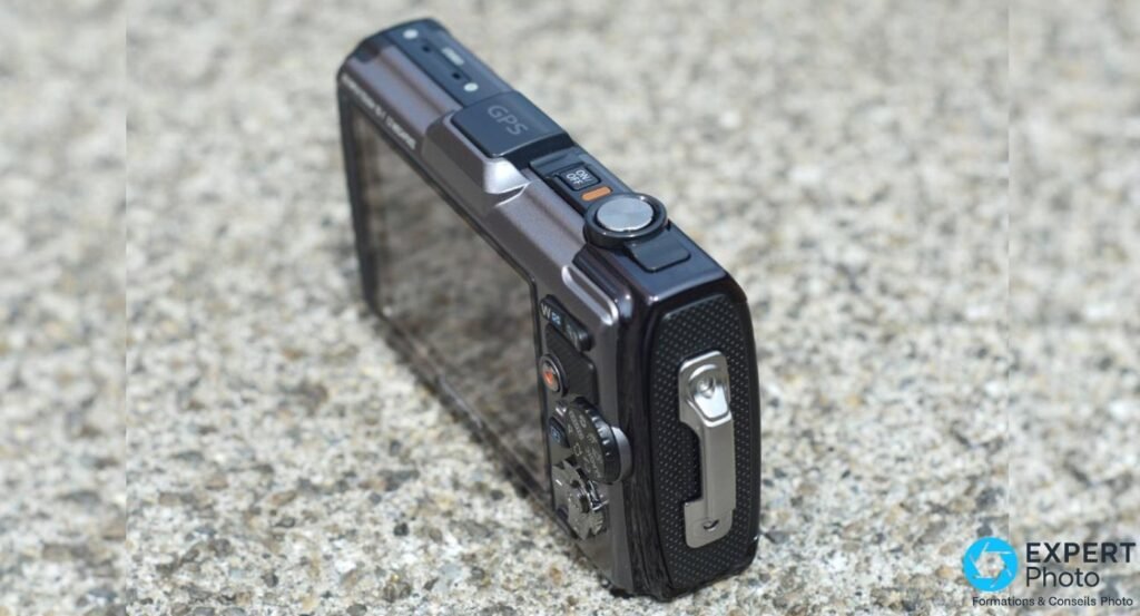 Olympus Tough TG-1 vu en plongée 3/4 arrière : déclencheur, cache GPS, roue de mode et attache dragonne visibles. 