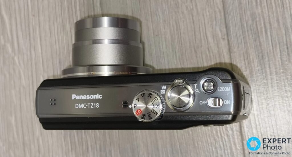Panasonic DMC-TZ18 vu de dessus, zoom déployé, molette modes P/A/S/M, bouton déclencheur et interrupteur ON/OFF visibles