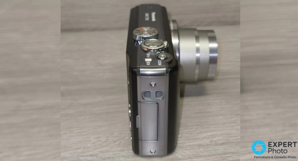 Panasonic DMC-TZ18 de profil gauche, zoom optique déployé sur plusieurs bagues argentées, fond bois flou