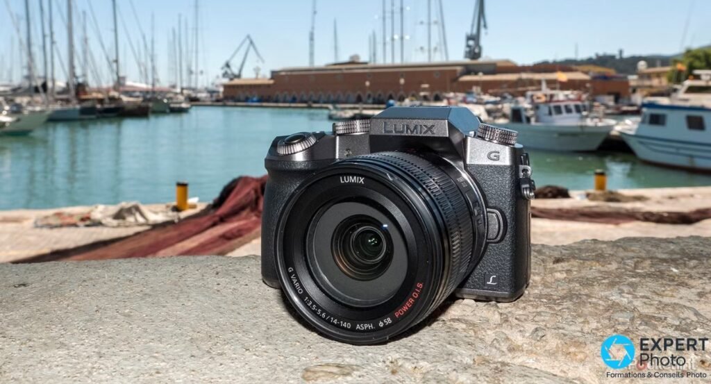 Panasonic Lumix G7 face avant posé sur un parapet en pierre, port maritime avec voiliers et halle de chantier naval en fond.