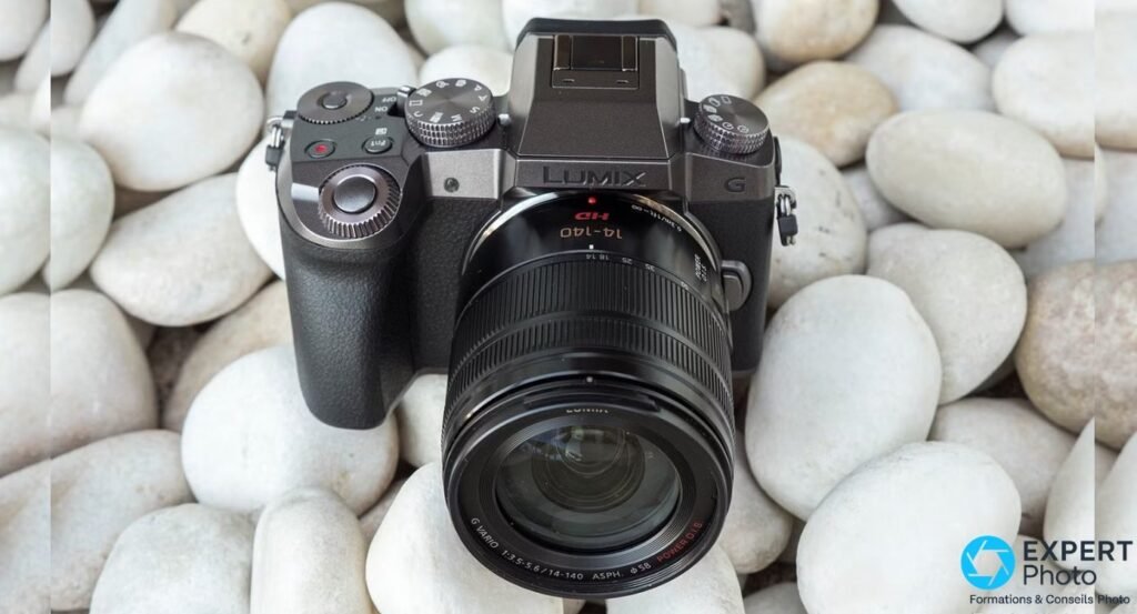 Panasonic Lumix G7 noir vu de face avant, objectif Lumix G Vario 14-140 mm OIS monté, posé sur fond de galets clairs.