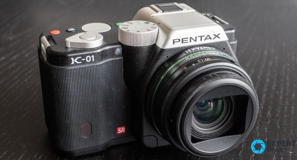 Pentax K-01 noir et argent de 3/4 avant gauche, objectif à bande verte monté avec pare-soleil noir, logo K-01 lisible