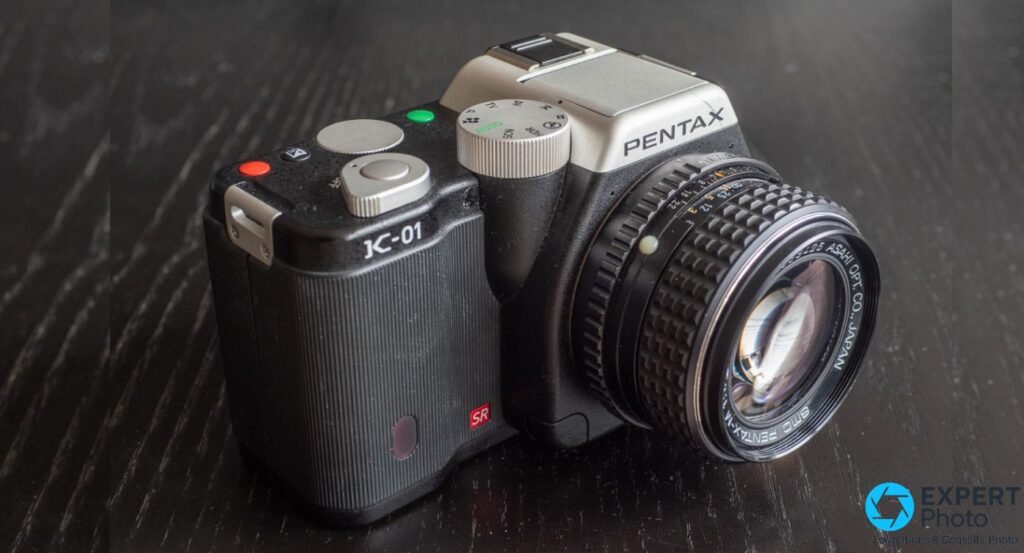 Pentax K-01 noir/argent 3/4 avant droit, objectif vintage SMC Pentax-M monté, inscription Asahi Opt Co Japan lisible