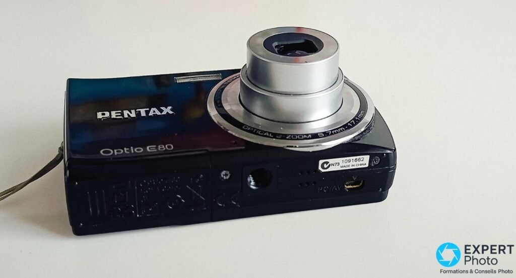Pentax Optio E80 noir vu de trois quarts arrière, dessous avec filetage trépied, port PC/AV mini-USB et étiquette.