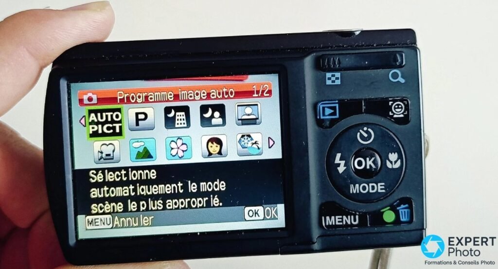 Pentax Optio E80 tenu à la main, écran arrière affichant le menu Programme image auto avec icônes de modes scènes. 