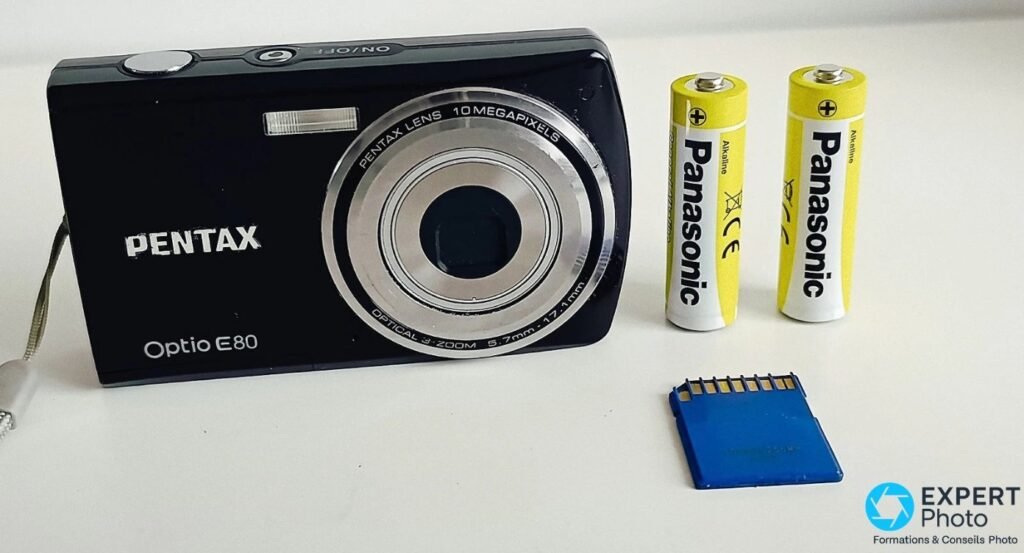  Pentax Optio E80 noir vu de face, objectif rétracté, avec deux piles AA Panasonic Alkaline et une carte SD bleue à côté. 
