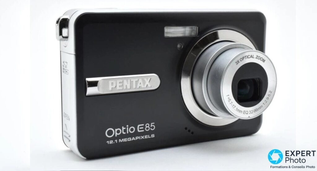 Pentax Optio E85 noir en vue trois quarts avant, marquage OPEN visible sur la tranche latérale gauche, zoom déployé.