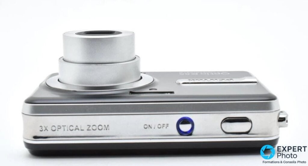 Pentax Optio E85 vu de dessus, zoom 3X OPTICAL ZOOM déployé en deux étages, bouton ON/OFF cerclé de bleu et déclencheur.