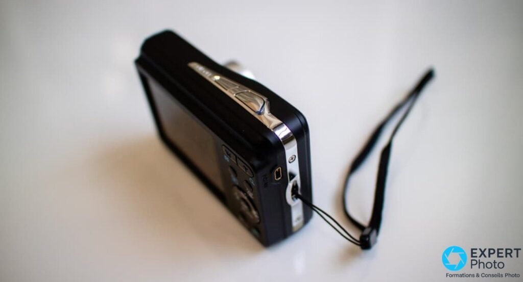 Pentax Optio E90 vu de trois-quarts arrière, déclencheur chromé visible et cordon noir tressé déployé en boucle. 