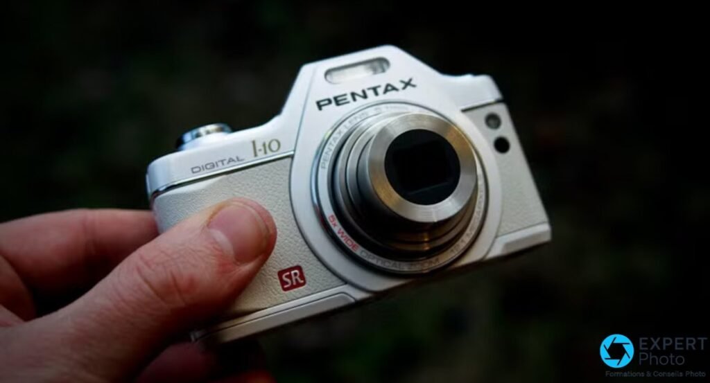 Pentax Optio I-10 blanc tenu en main, objectif zoom 5x déployé, étiquette SR rouge visible.