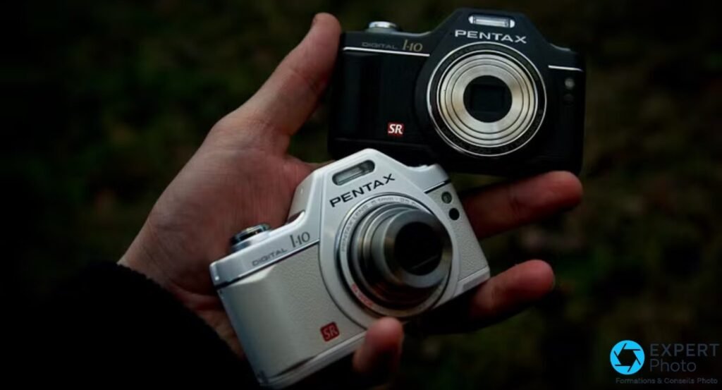 Deux Pentax Optio I-10 dans une main, modèle noir au fond et blanc devant, objectifs rétractés.