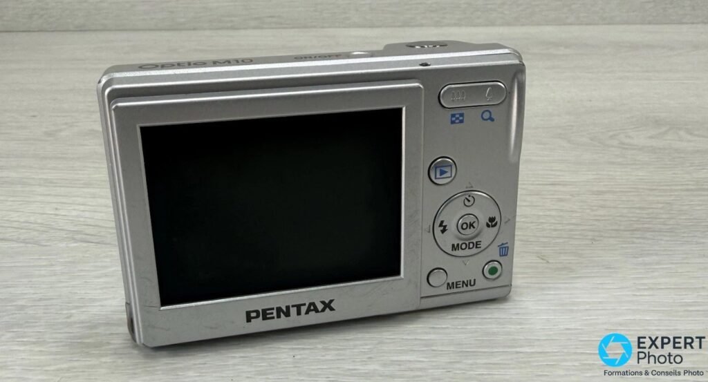 Pentax Optio M10 vu de l'arrière, écran LCD 2,5 pouces éteint, molette directionnelle, boutons MENU et MODE.