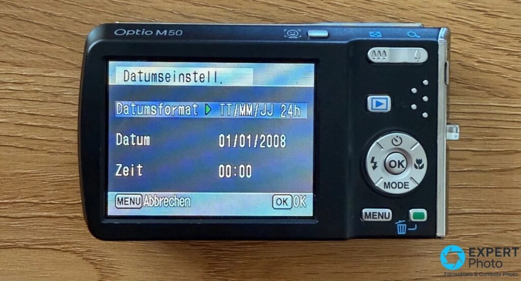 Pentax Optio M50 vu de l'arrière, écran allumé sur le menu de réglage de date (01/01/2008) affiché en allemand. 