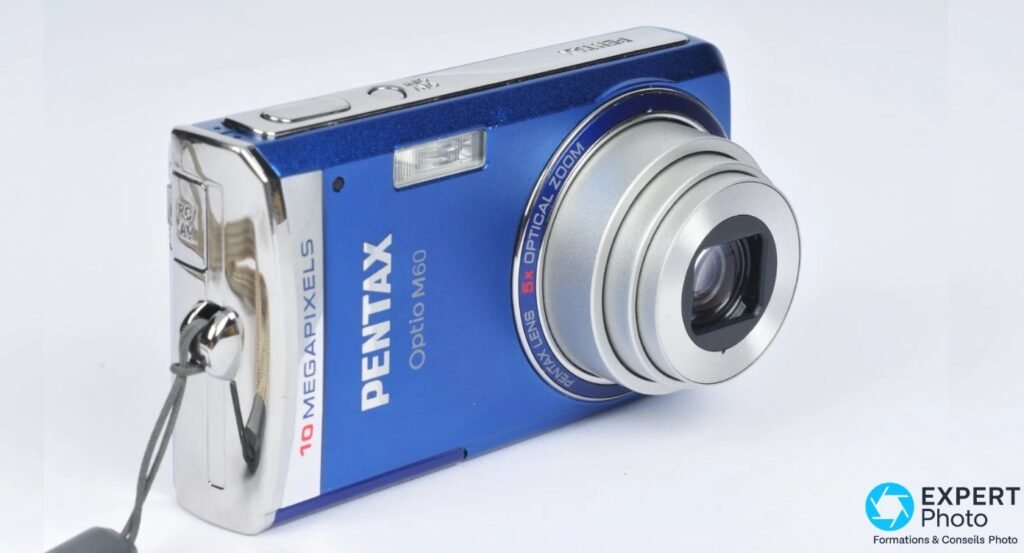 Pentax Optio M60 bleu, 3/4 avant-gauche, dragonne attachée, objectif étendu, prise RC/AV visible sur la tranche