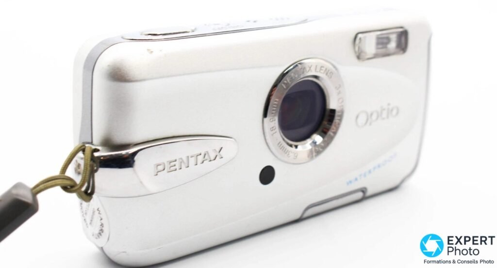 Pentax Optio W30 argenté, face avant en contre-plongée, logo PENTAX et objectif visibles, cordelette kaki attachée