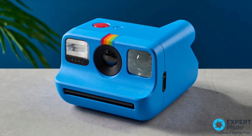  Polaroid Go Gen 2 bleu posé sur béton gris, vue 3/4 avant, fond bleu foncé avec feuille de palmier floue