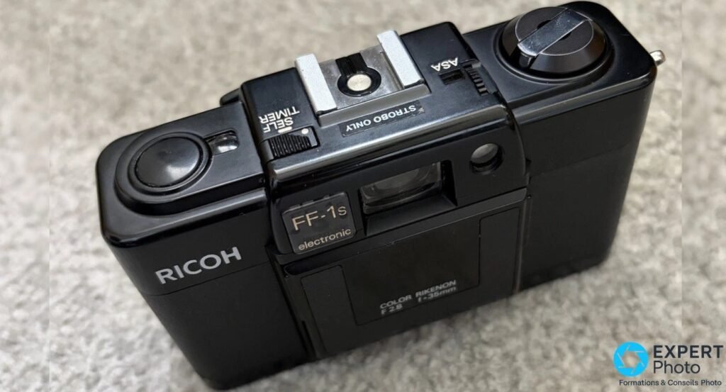 Ricoh FF-1S vu en trois-quarts arrière, capot avec SELF TIMER, STROBO ONLY et ASA, déclencheur et manivelle visibles