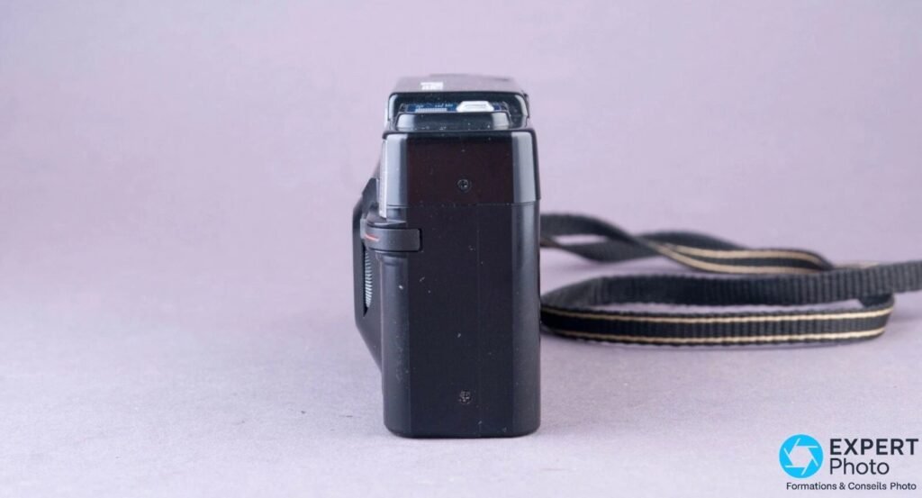 Ricoh FF-3 AF Super vu de profil droit sur fond gris, dragonne noire à bandes claires posée derrière l'appareil.