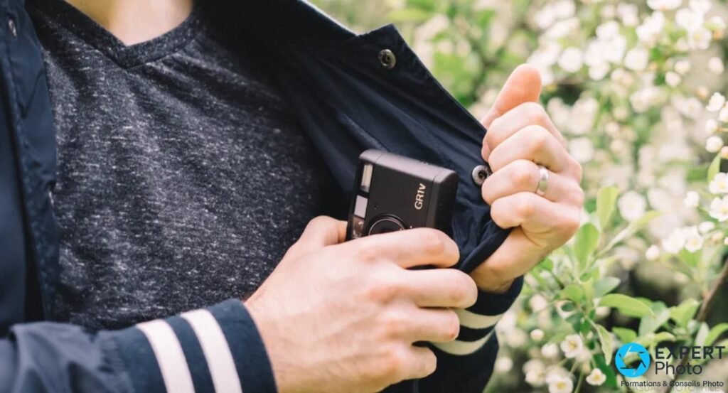 Main rangeant un Ricoh GR1v dans une veste, fond exterieur fleuri illustrant un usage compact en voyage leger. 