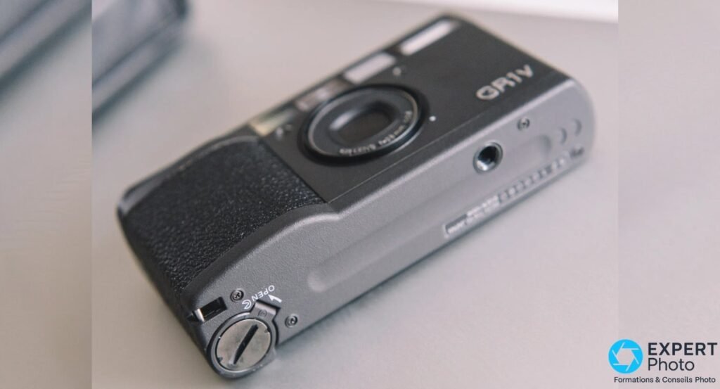 Ricoh GR1v vu de dessous en angle, trappe de compartiment pile marquee OPEN et pas de vis trepied visibles.