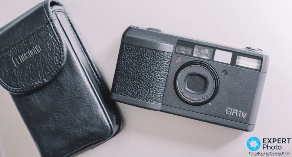 Ricoh GR1v noir pose a plat a cote de son etui d'origine en cuir noir marque RICOH, sur fond clair.