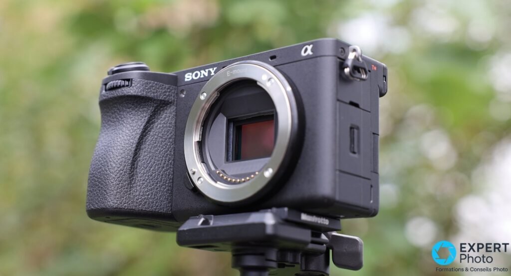 Sony A6700 sans objectif sur trépied Manfrotto, vu de 3/4 avant gauche, monture E et capteur visibles, fond végétal flou.