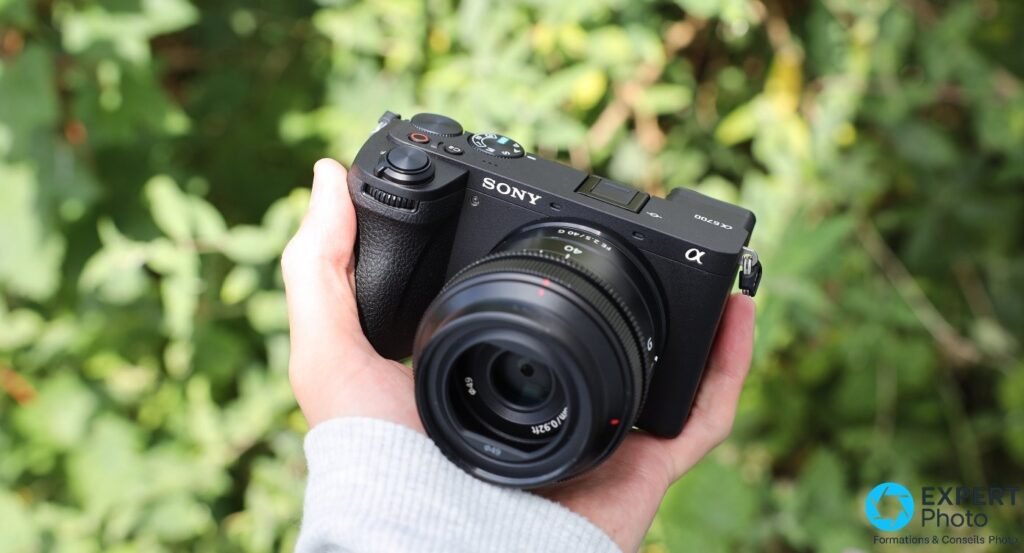Sony A6700 avec FE 2.5/40 G tenu en main, vue 3/4 avant gauche, fond de feuillage vert flou.