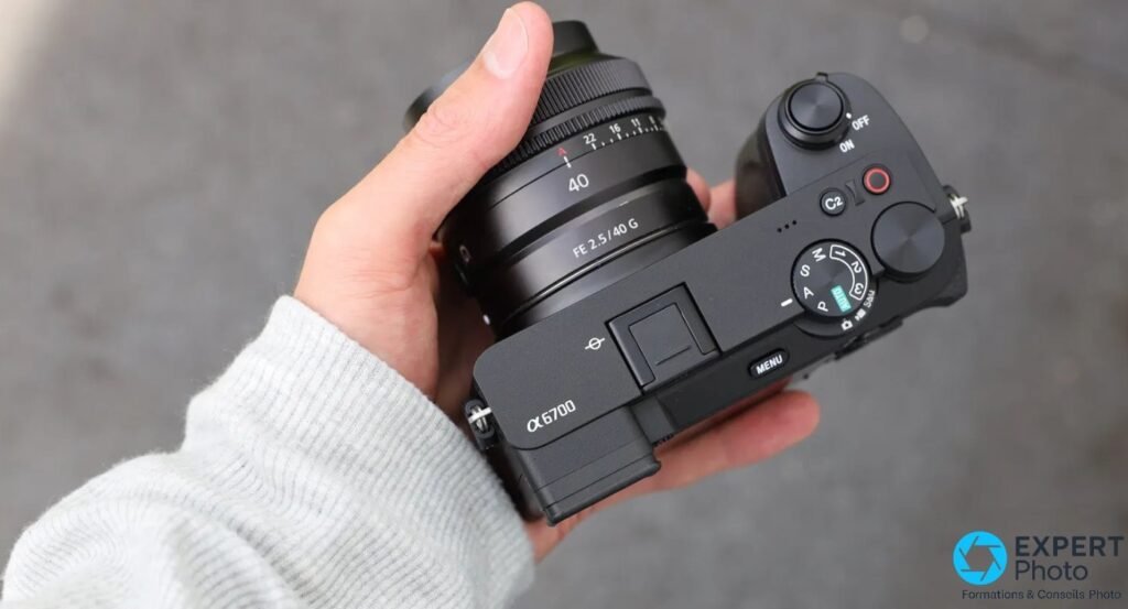 Sony A6700 tenu en main, vue de dessus avec FE 2.5/40 G, dial PASM, MENU, ON/OFF et α6700 visibles.