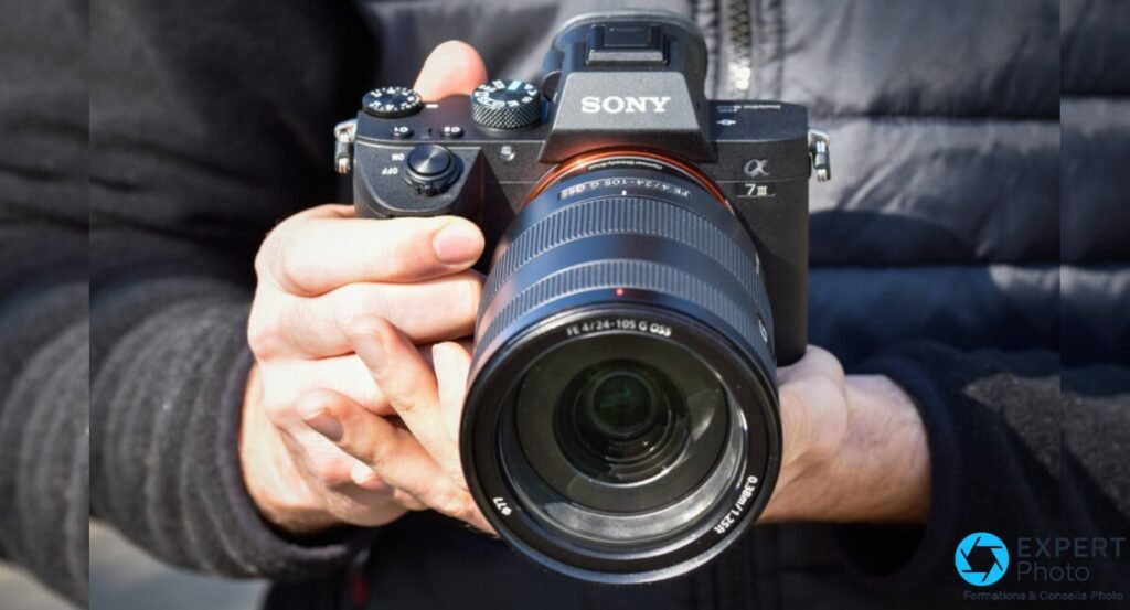 Sony A7 III avec objectif FE 24-105 G OSS au premier plan, logo G et pastille rouge visibles, boîtier tenu à deux mains