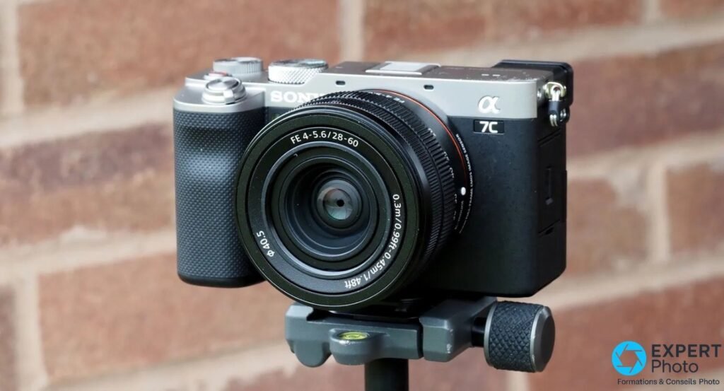Sony A7C avec objectif FE 28-60 mm monté sur tête de trépied, fond mur de briques rouges flou.