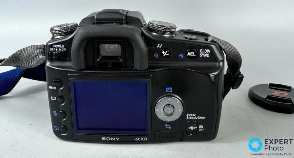 Vue arrière du Sony Alpha 100 (DSLR-A100) avec marquage α 100, bouton Super SteadyShot ON/OFF et bouchon d'objectif.