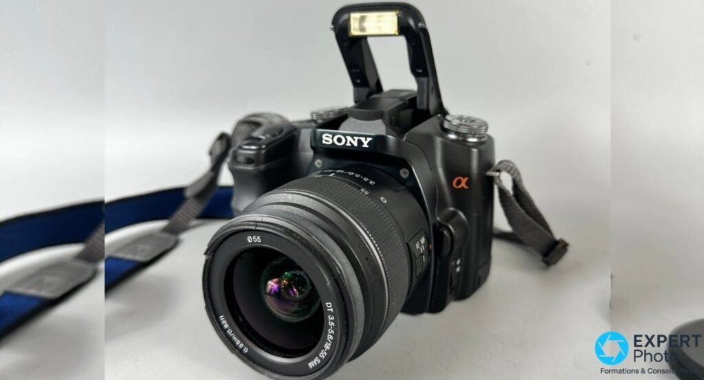 Vue de face du Sony Alpha 100 (DSLR-A100) avec flash pop-up intégré déployé et objectif 18-55 mm SAM monté.