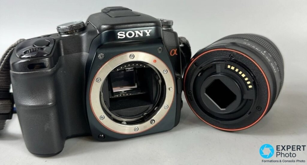 Sony Alpha 100 boîtier nu avec monture A ouverte montrant miroir et contacts, objectif Sony posé à côté contacts visibles.
