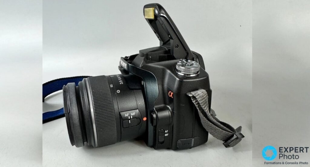 Profil gauche du Sony Alpha 100 avec flash intégré déployé, sélecteur AF/MF du boîtier et objectif 18-55 mm SAM visible.